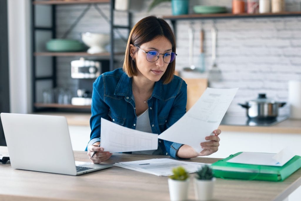 young woman working with computer while consulting some invoices and documents in the kitchen